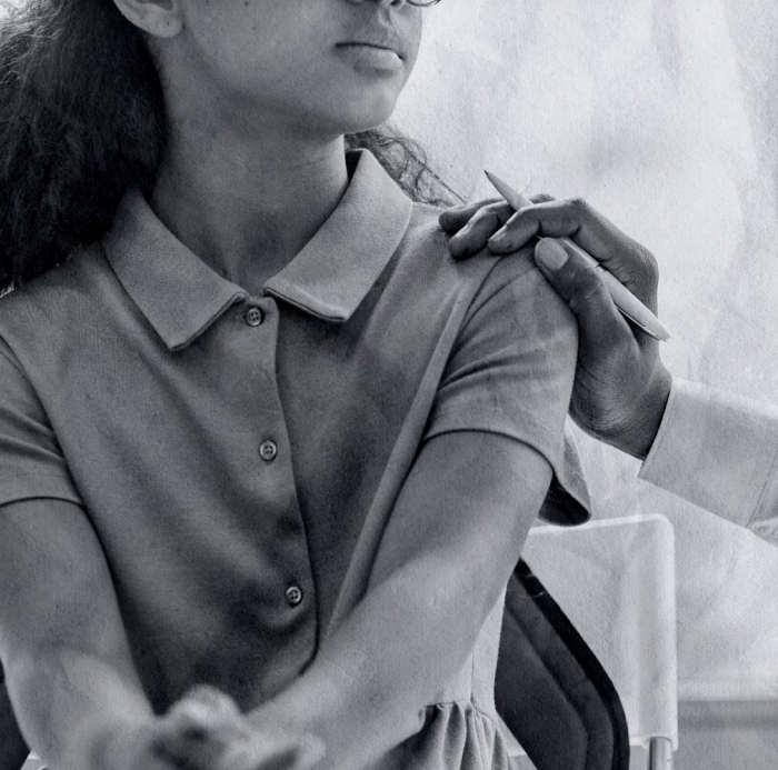 A healthcare professional from an équipe mobile administers a vaccination shot in the upper arm of a young adolescent girl who is seated, wearing a short-sleeved button-up dress.